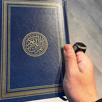 Hand holding blue Quran with gold Arabic calligraphy and ornate border, ideal for Islamic study and devotion.